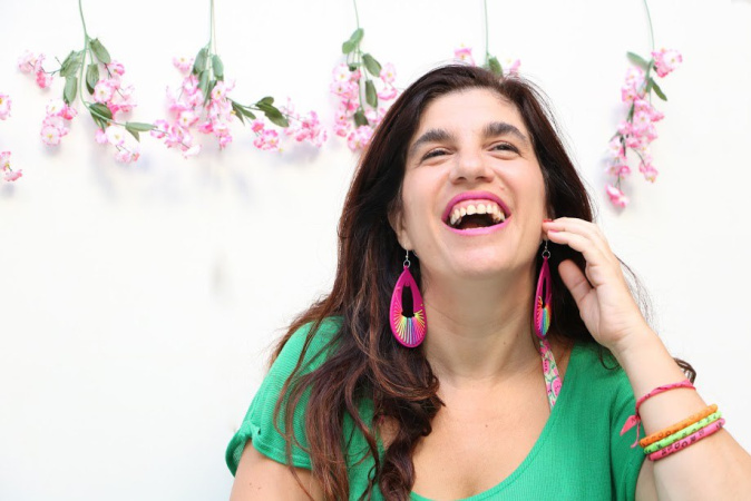 Luciana Peker se retrata en una foto. Es una mujer de cabello cafe obscuro, y largo, más abajo de sus hombros.  Está sonriendo con vista hacía al horizonte y arriba, mano levantada a la mejilla de su cara, con sonrisa grande y boca abierta. Viste una camiseta verde con aretes grandes y pulseras de muchos colores. En el fondo contra pared hay seis ramitas de flores color rosa pálida con hojas verdes, colgadas cada rama de manera deocrativa, hojas y pétalos hacía abajo.